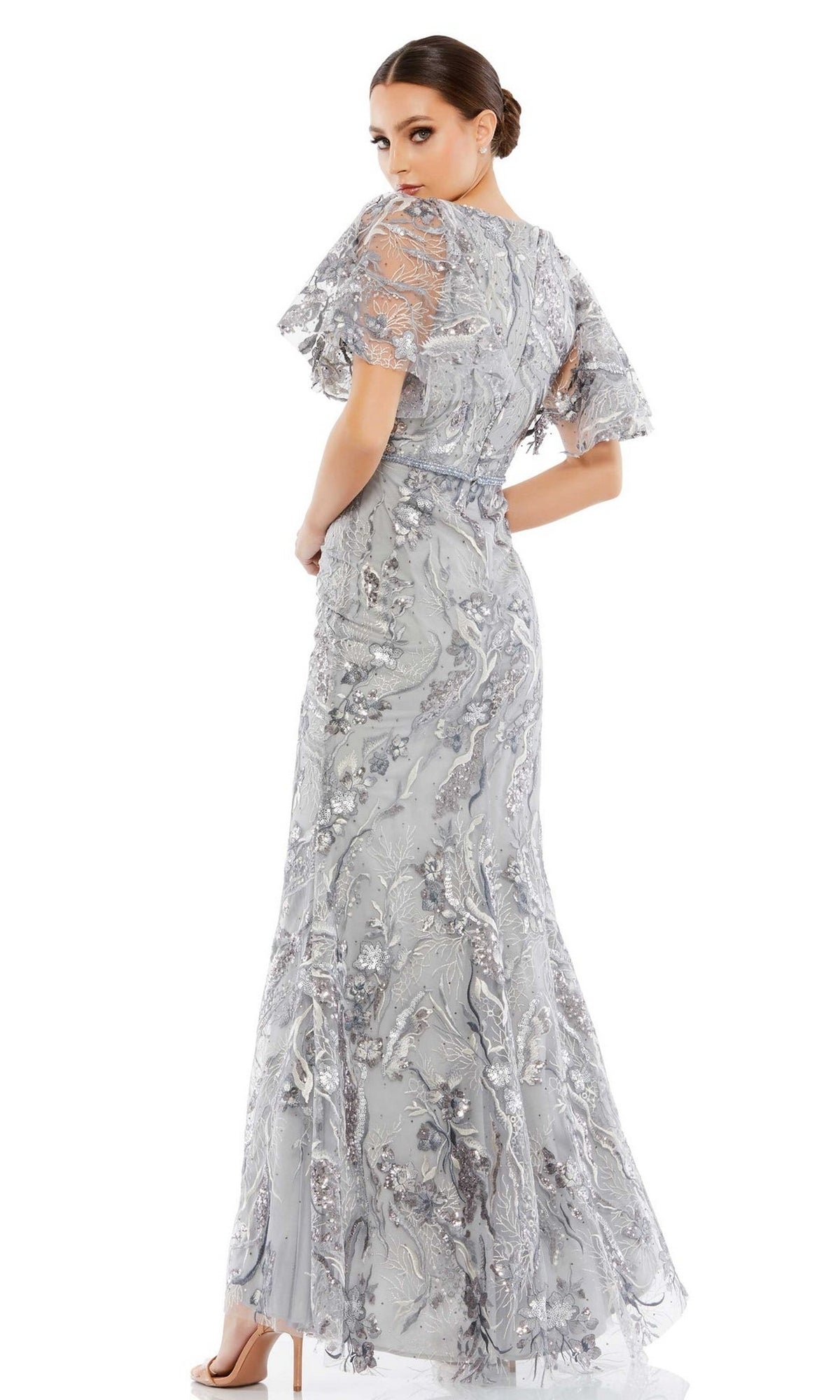 A woman in the Long Formal Dress 67908 by Mac Duggal, featuring a platinum fitted silhouette with sheer, embellished flutter sleeves and floral embroidery, stands in high heels and glances over her shoulder.